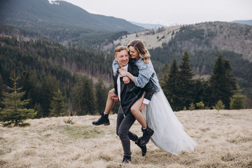 Stylish couple in the autumn mountains. A guy in a leather jacket and a young girl in a gray blue wedding dress and a denim jacket having fun together on the background of a sky, forest and landscapes