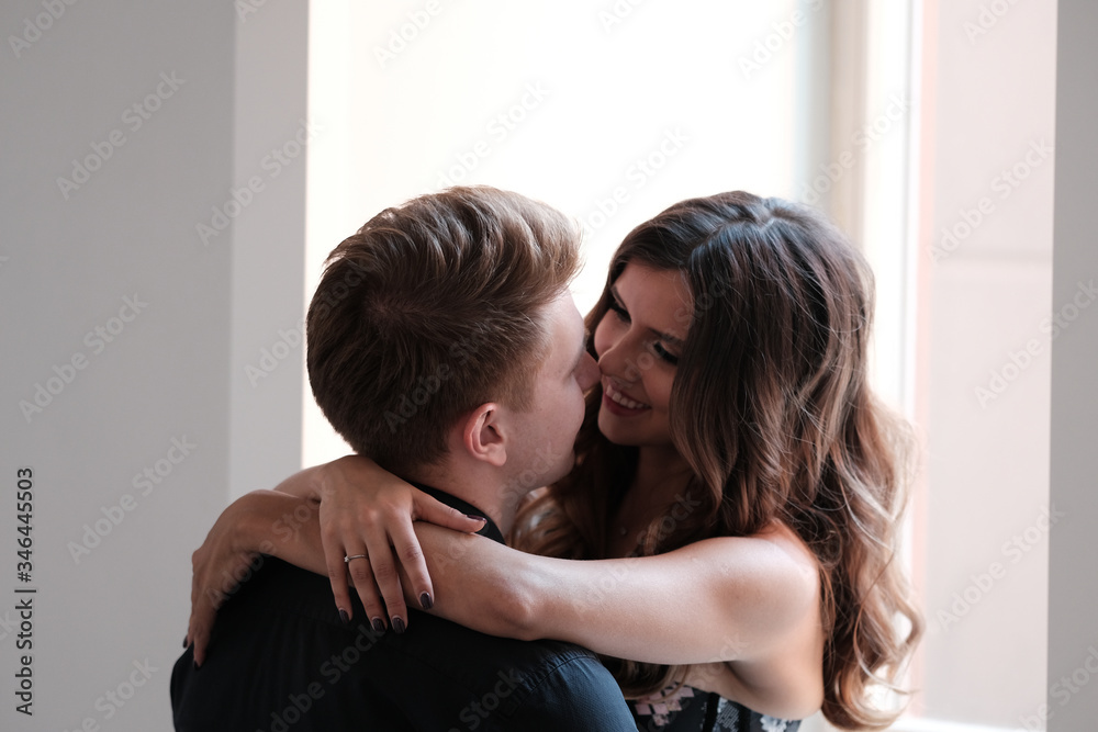 A young couple - a girl in a magnificent dress and a guy in a black shirt and trousers hug and kiss in a hotel room. A young couple - a girl in a magnificent dress and a guy in a black shirt and trousers hug and kiss in a hotel room.