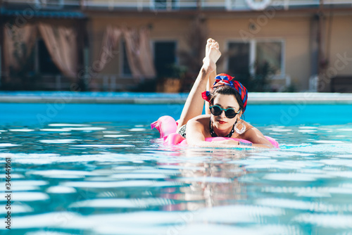  Beautiful girl by the pool with long dark hair and make-up resting at a spa resort. In a black retro bathing suit, scarf, hat and glasses, smiles sunbathes on a pink inflatable mattress in the motel