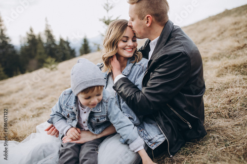 Stylish family in the autumn mountains. A guy in a leather jacket and a young girl in a gray-blue wedding dress with their son sitting on the grass together on the background of forest and landscapes