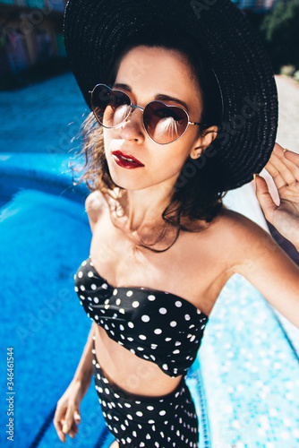 Beautiful girl by the pool with long dark hair and make-up resting at a spa resort. In a black retro bathing suit, scarf, hat and glasses, smiles sunbathes on a pink  inflatable mattress in the motel © сергей макаров