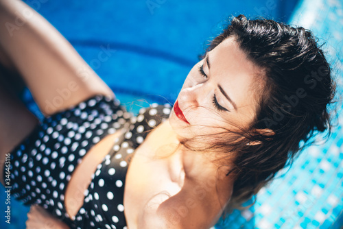 
Beautiful girl by the pool with long dark hair and make-up resting at a spa resort. In a black retro bathing suit, scarf, hat and glasses, smiles sunbathes on a pink 
inflatable mattress in the motel