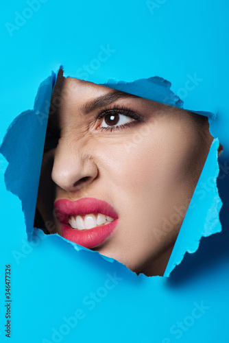 Aggressive woman with pink lips looking across ripped blue paper © LIGHTFIELD STUDIOS
