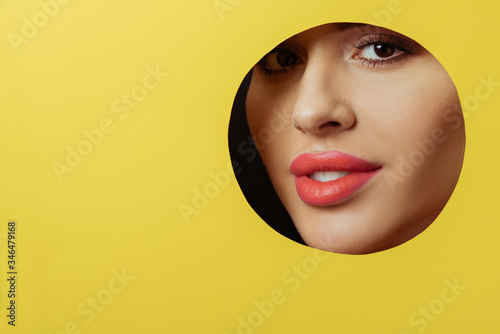 Woman with coral lips and smoky eyes looking at camera and smiling across round hole in yellow paper on black © LIGHTFIELD STUDIOS