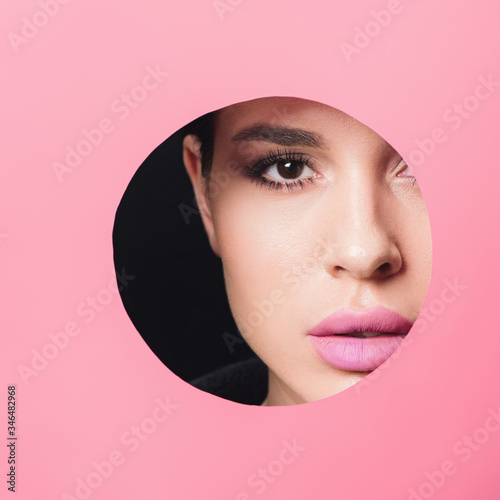Beautiful girl with smoky eyes and pink lips looking at camera across hole in paper on black background © LIGHTFIELD STUDIOS