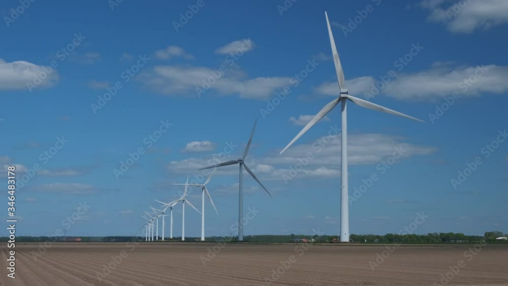 Row of wind turbines in an agricultural environment with a blue sky and fluffy clouds above during a sunny springtime day. Row of wind turbines in an agricultural environment with a blue sky and fluffy clouds above during a sunny springtime day.