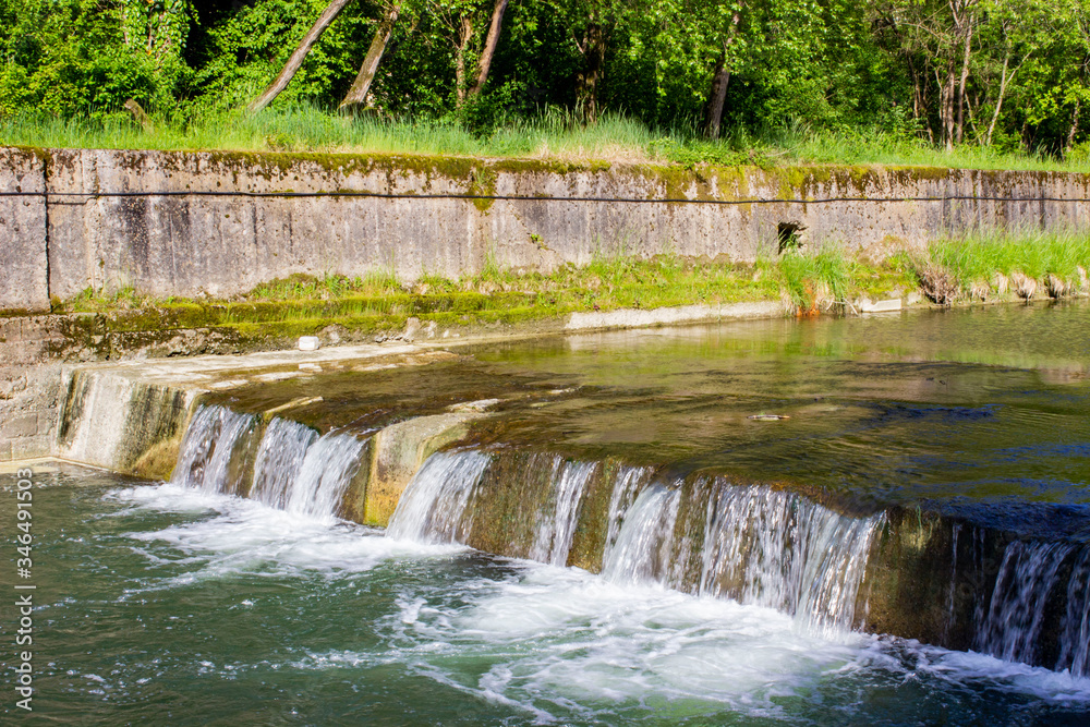 Spring landscape. The threshold of a mountain meandering river enclosed in stone fences. The water drops rapidly. Green plants, shrubs, and trees. Sunny weather. Spring landscape. The threshold of a mountain meandering river enclosed in stone fences. The water drops rapidly. Green plants, shrubs, and trees. Sunny weather.