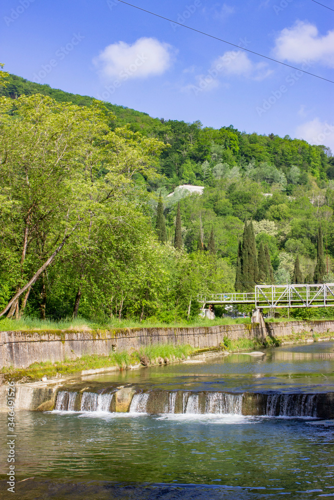 Spring landscape. The threshold of a mountain meandering river enclosed in stone fences. The water drops rapidly. Green plants, shrubs, and trees. Sunny weather.