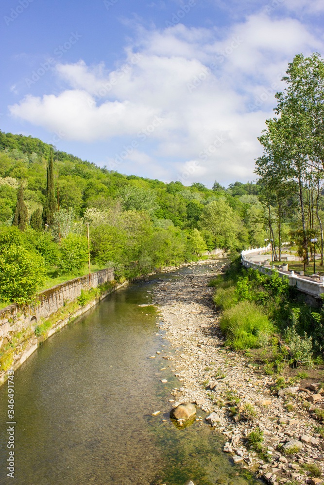 Spring landscape. A mountain meandering river enclosed in stone fences. Shallow, with a bare stone bottom on the side. Green plants, shrubs, and trees. Sunny weather. Spring landscape. A mountain meandering river enclosed in stone fences. Shallow, with a bare stone bottom on the side. Green plants, shrubs, and trees. Sunny weather.