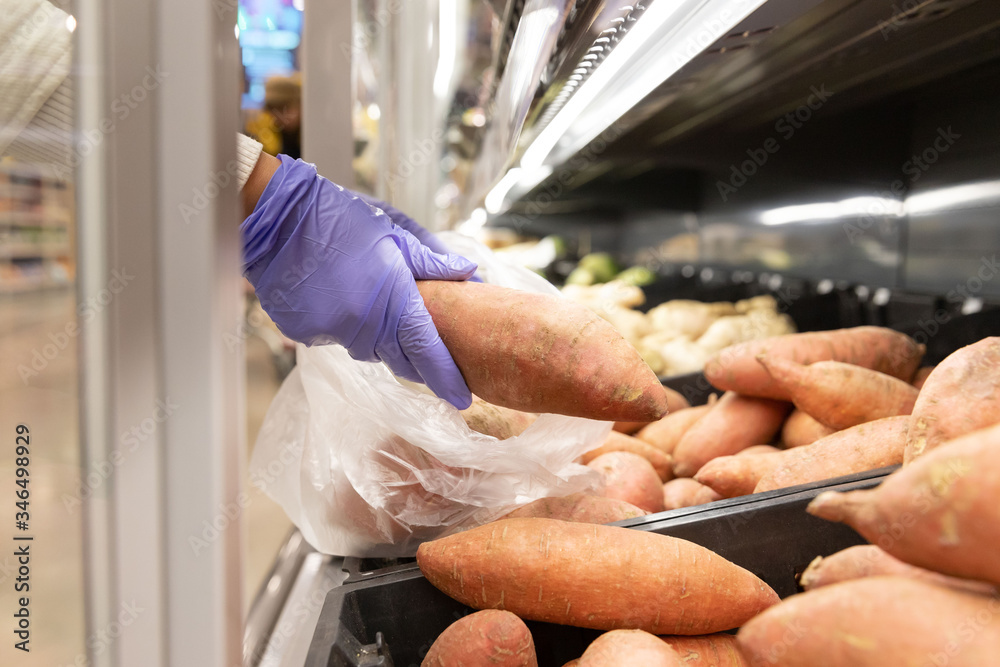Woman wears rubber protective medical gloves, chooses sweet potato folding in disposable plastic bag in supermarket, close up. Protective measures against coronavirus pandemic, covid-19 outbreak. 