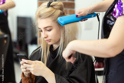 Beautiful blonde girl with long straight hair sits in a chair in a beauty salon. A professional hairdresser makes a girl curling after dyeing hair. The girl uses the phone during the procedure.