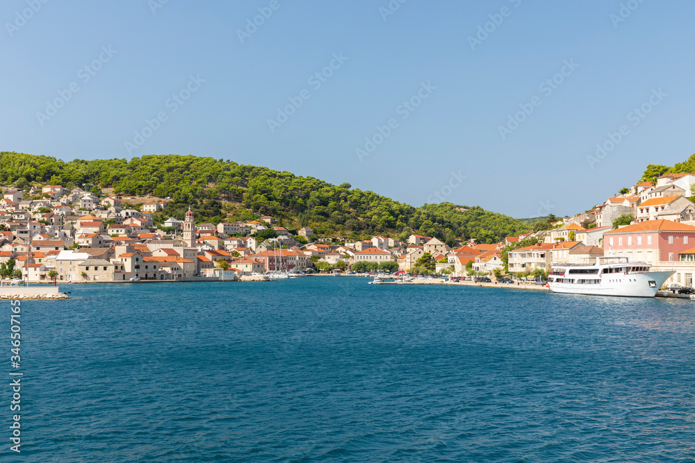 Pucisca town at Brac in Croatia, view from the sea on a sunny day and a blue sky in the summer. The port with it’s famous limestone from the island and a cruise ship. Idyllic place, beautiful scenery