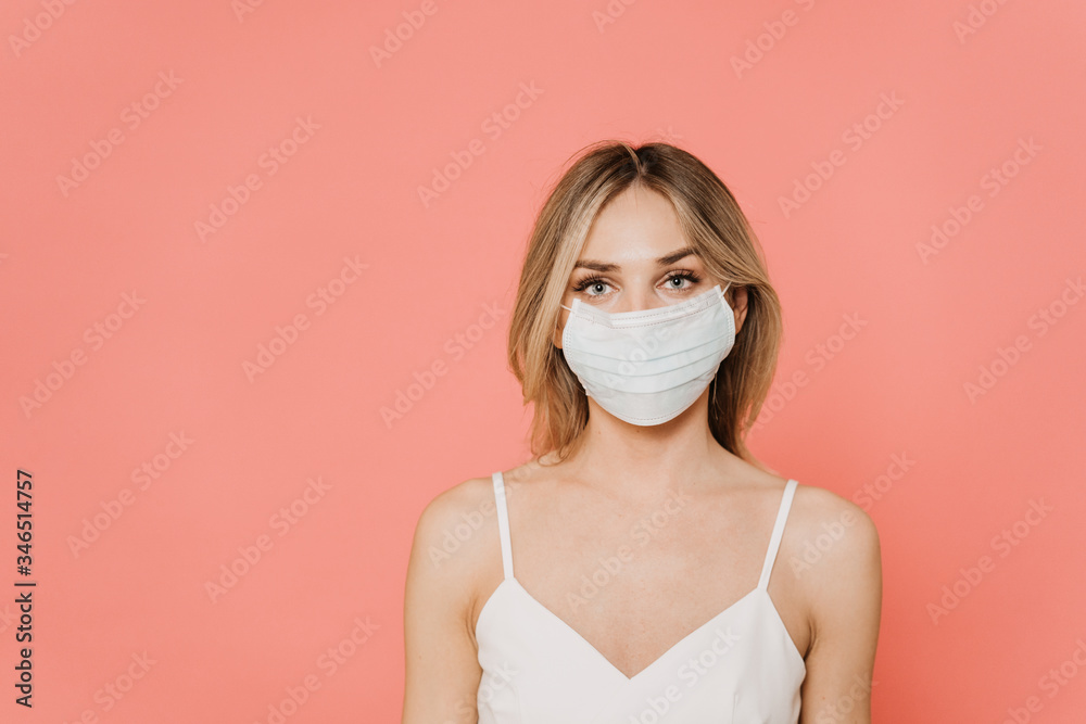 Beautiful blonde in surgical protective mask looking confident, waiting of the end of coronavirus pandemic at home, stands over pink background with copy space. Health care concept.