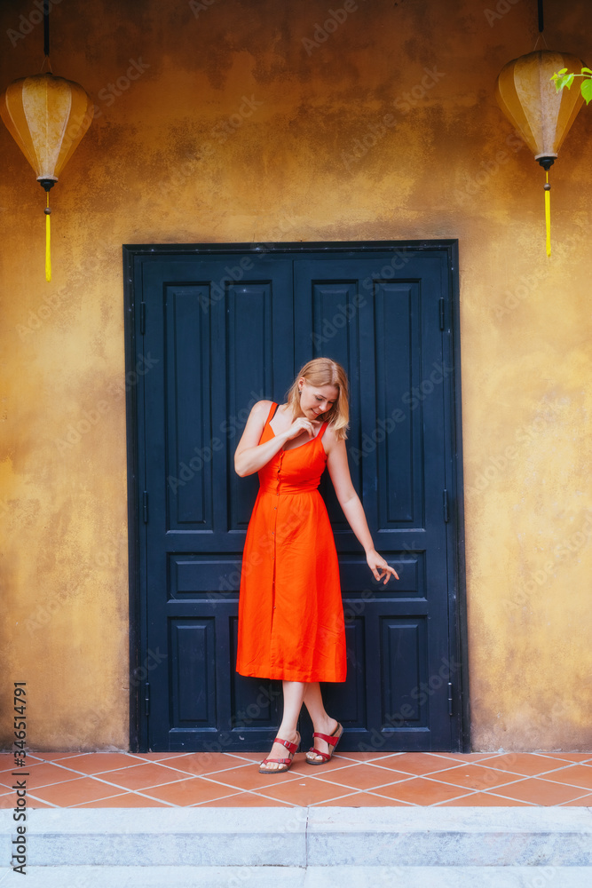 A beautiful girl in a red dress stands against the dark door of an old yellow house with Chinese lanterns.architecture of the ancient city of Hoi an .Vietnam.