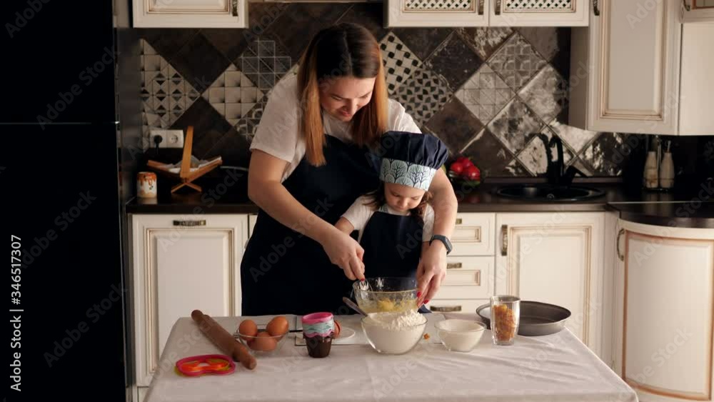 A little funny girl of three years helps her mother to cook in the kitchen. Mother and daughter in matching aprons prepare cookie dough together at home in a modern kitchen.