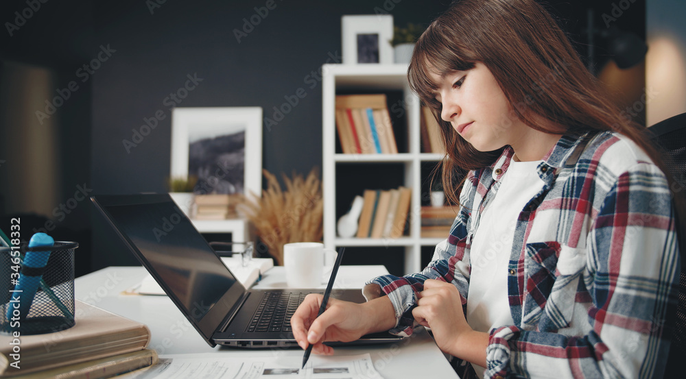 Side view of teen girl sitting at desk with workbook and laptop doing hometask, remote education Side view of teen girl sitting at desk with workbook and laptop doing hometask, remote education