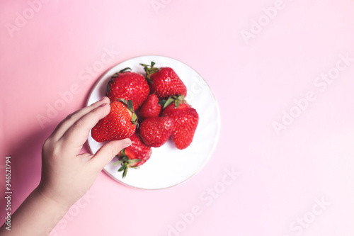 Top view Childs hand holding a strawberry on pink background, plate of strawberries. Summer healthy eating concept.