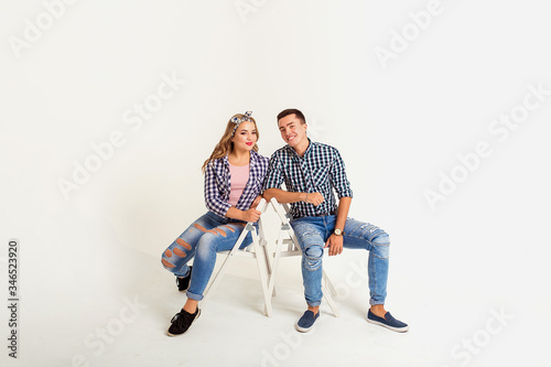 A beautiful girl and a manly guy of European appearance are sitting on chairs and looking at the camera, they are smiling, dressed stylishly in plaid shirts and jeans, on a white background, isolated