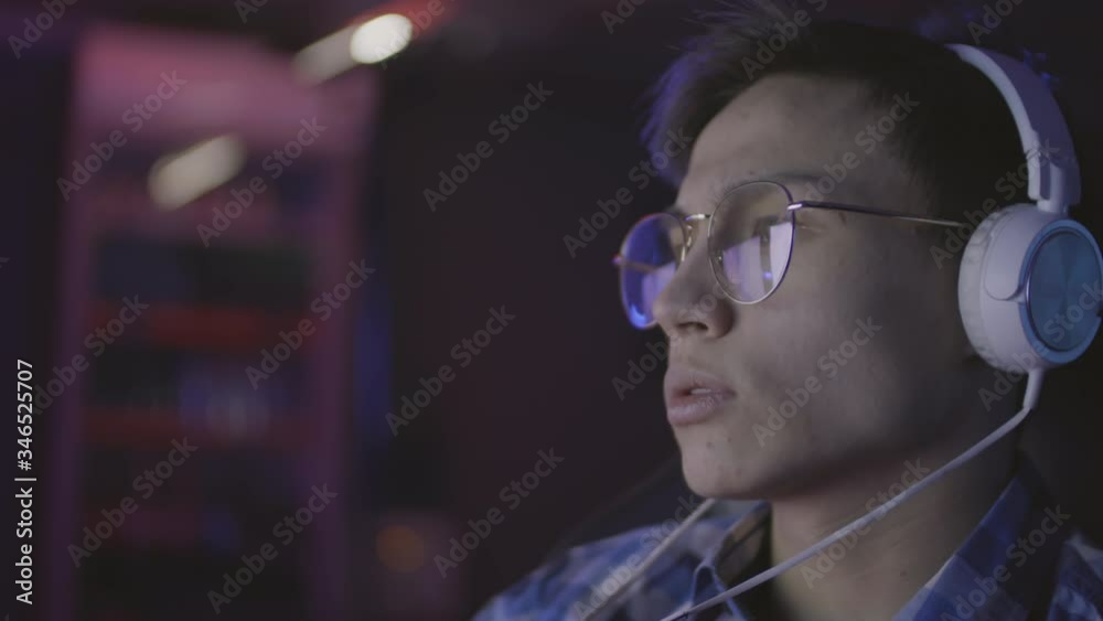 Close up portrait of young Asian man wearing blue plaid shirt, eyeglasses and headphones sitting at computer and streaming Close up portrait of young Asian man wearing blue plaid shirt, eyeglasses and headphones sitting at computer and streaming