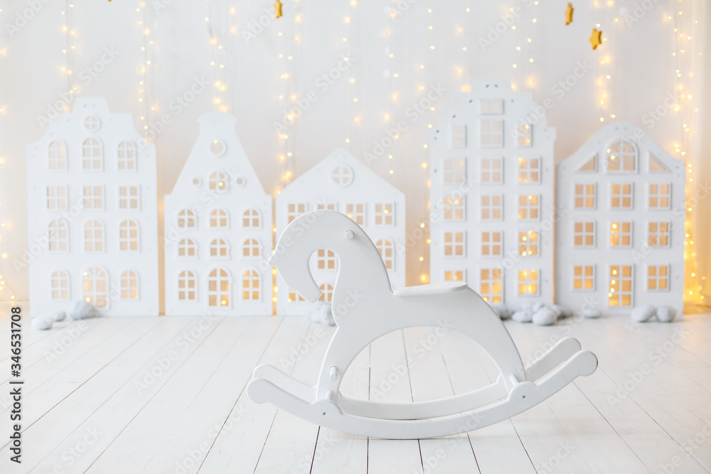 White children room with decorative wooden houses. White wooden toy rocking horse on wall background. Toy houses in Christmas decor. Interior of the children's room. Paper garland in the form of stars
