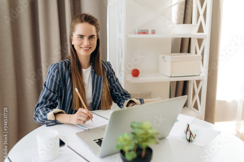 Distant work. Attractive smiling girl in a stylish formal clothes works from home sitting on her workplace and using a laptop