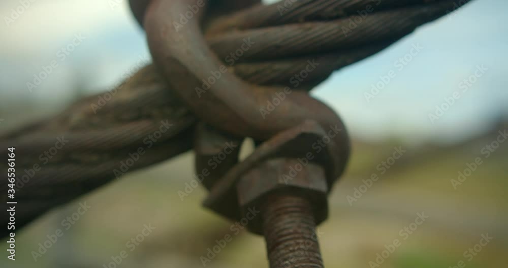 Old rusty coarse thread bolt with huge hooked nut secures woven steel cable of suspension pedestrian bridge against blurred background of grass and sky. Close-up The wire rope divides frame diagonally Old rusty coarse thread bolt with huge hooked nut secures woven steel cable of suspension pedestrian bridge against blurred background of grass and sky. Close-up The wire rope divides frame diagonally