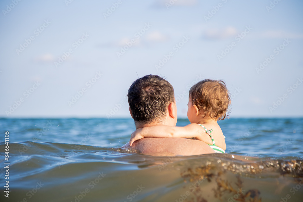 family vacation concept. dad holds a little cute toddler daughter in the sea water and watching the horizon back view family vacation concept. dad holds a little cute toddler daughter in the sea water and watching the horizon back view