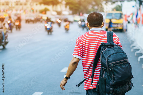 Back view of male patient with mask in red and white shirt with a black backpack standing at bus stop and waving his hand for Taxi or Bus in the city to go to the hospital.