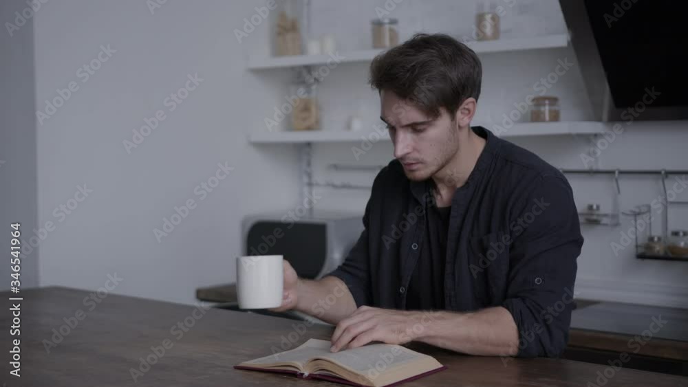 Young man sitting at a table reading a book and drinking a drink from a white mug, smiling, concept of spending time at home, leisure, isolation Young man sitting at a table reading a book and drinking a drink from a white mug, smiling, concept of spending time at home, leisure, isolation