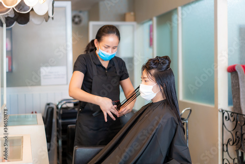 Asian young woman wearing face masks to protect themselves from Covid-19 during hairdresser trimming black hair with scissors in beauty salon.