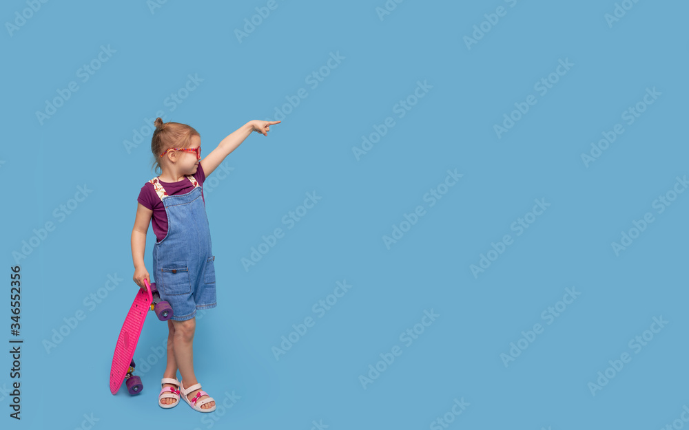Cool little toddler girl in bright clothes sits on a skateboard and looking at the camera isolated on blue background Cool little toddler girl in bright clothes sits on a skateboard and looking at the camera isolated on blue background