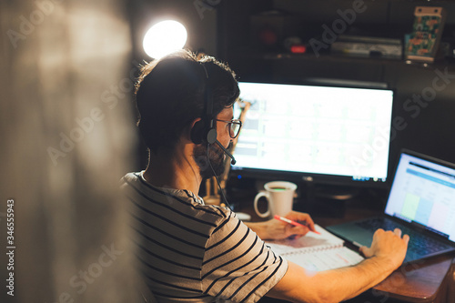 Modern young man working remotely from home at night. Casual millennial guy typing on pc notebook surfing internet looking at screen enjoying distant job