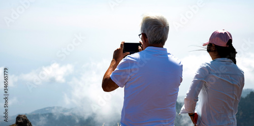 View from the top of the mountain. Tourists: an elderly man and woman stand on the top of a mountain and take photos on their phone of the clouds under their feet. Copy space.