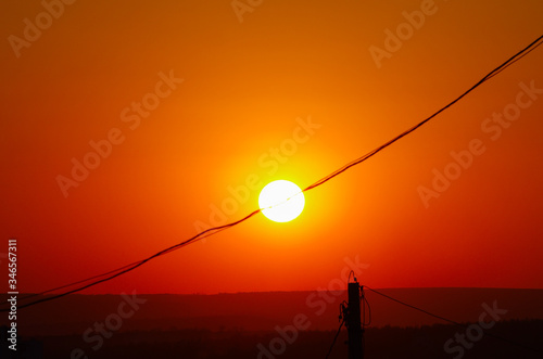 The disc of the sun crossed by an electricity line in orange sky at sunset golden hour