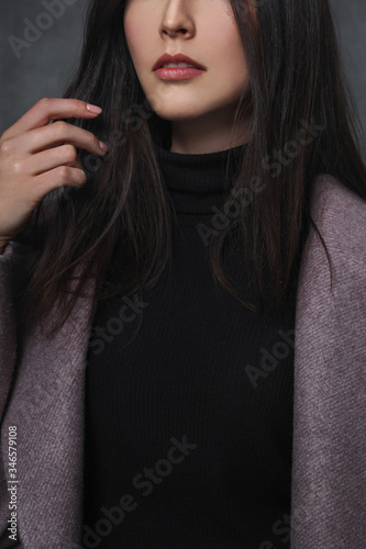 Beautiful young brunette girl in a black sweater and lilac coat on a gray background. Close-up, sensual lips and hands.