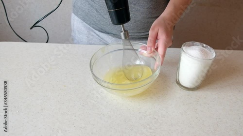 Woman with kitchen mixer whips egg whites and sprinkles sugar.  Electric mixer beats. Girls hand holds glass bowl. Chicken egg for tasty dish.