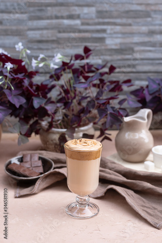 A popular Korean drink during self-isolation is Dalgona coffee. A delicious modern cold beverage with chocolate. Creamer, napkin is on the table. On a background is potted plants. Vertical still life.