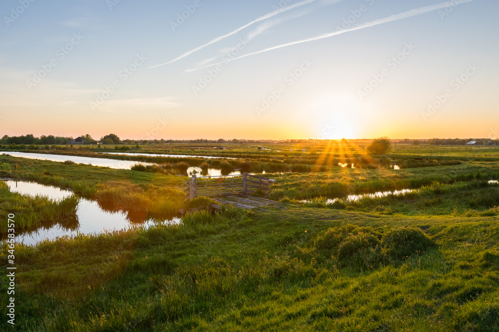 Sun sets over the wide open Dutch polder landscape. Green meadows intersected with ditches filled with water. - obrazy, fototapety, plakaty 