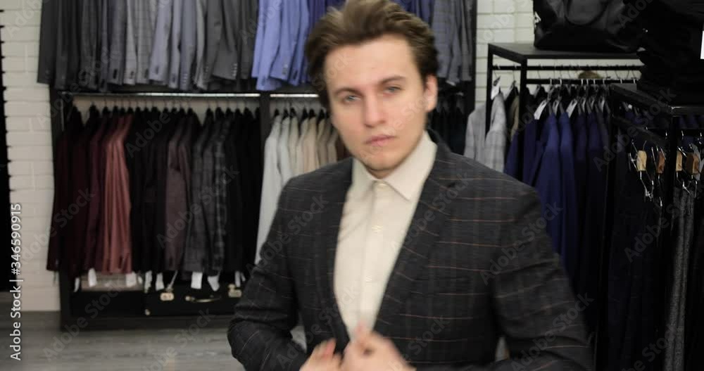 Young man is trying elegant suit with shirt in clothing menswear store in mall looking at camera as at mirror and smiling. Guy choosing buying costume. Portrait of man is fastening shirt, front view. Young man is trying elegant suit with shirt in clothing menswear store in mall looking at camera as at mirror and smiling. Guy choosing buying costume. Portrait of man is fastening shirt, front view.