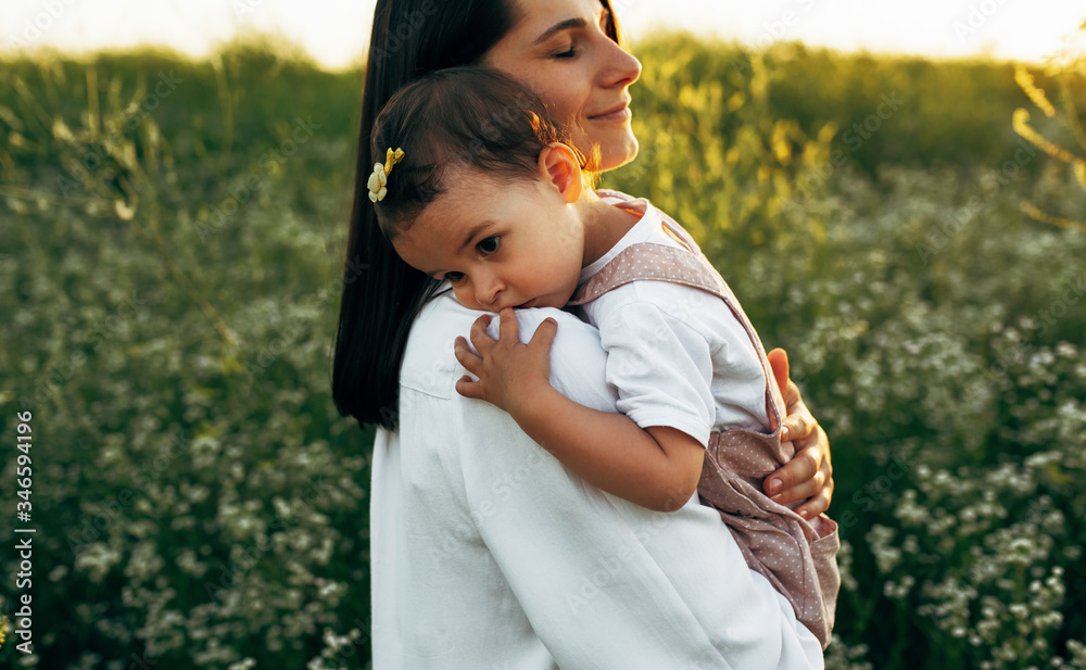 Closeup outdoor image of smiling mother and cute little girl embrace each other on the field on sunset sunlight. Mom and daughter walking on a flower field on nature background. Closeup outdoor image of smiling mother and cute little girl embrace each other on the field on sunset sunlight. Mom and daughter walking on a flower field on nature background.