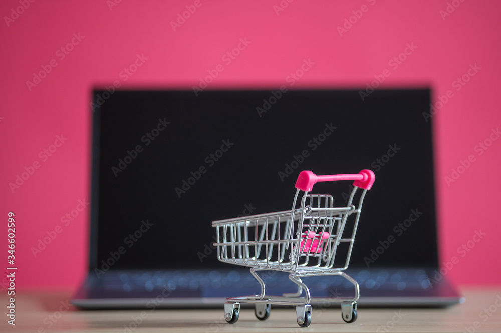 Online shopping concept. Mini grocery cart and laptop on a pink background. Small cart and personal computer.