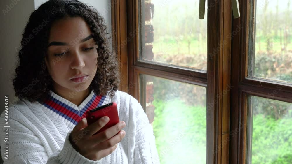 Sad beautiful mixed race African American girl teenager young woman during Coronavirus COVID-19 pandemic lockdown, sitting by a window using her mobile cell phone or smartphone for social media