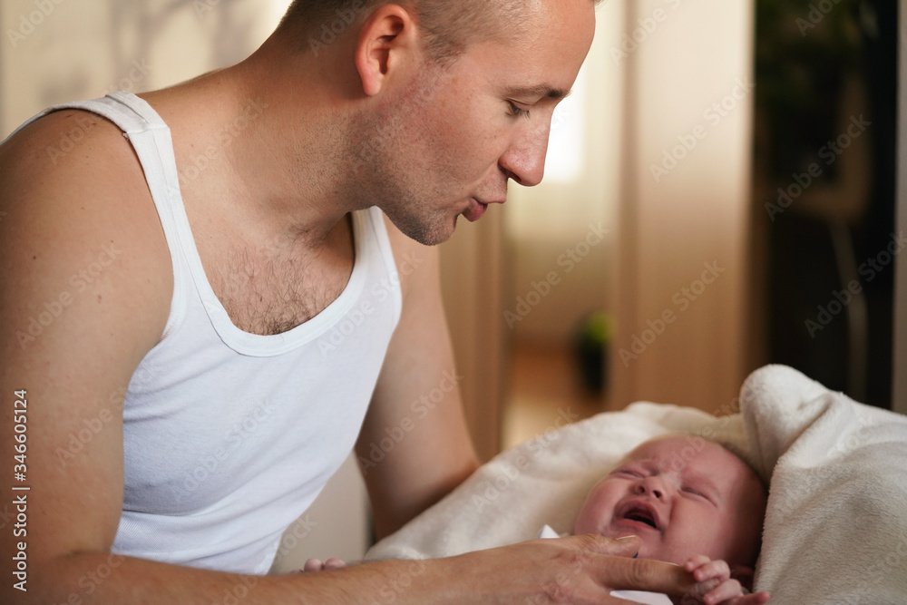 Young man wearing only vest top, playing with his baby son, trying to calm down, screaming kid as he holds him on hands Young man wearing only vest top, playing with his baby son, trying to calm down, screaming kid as he holds him on hands