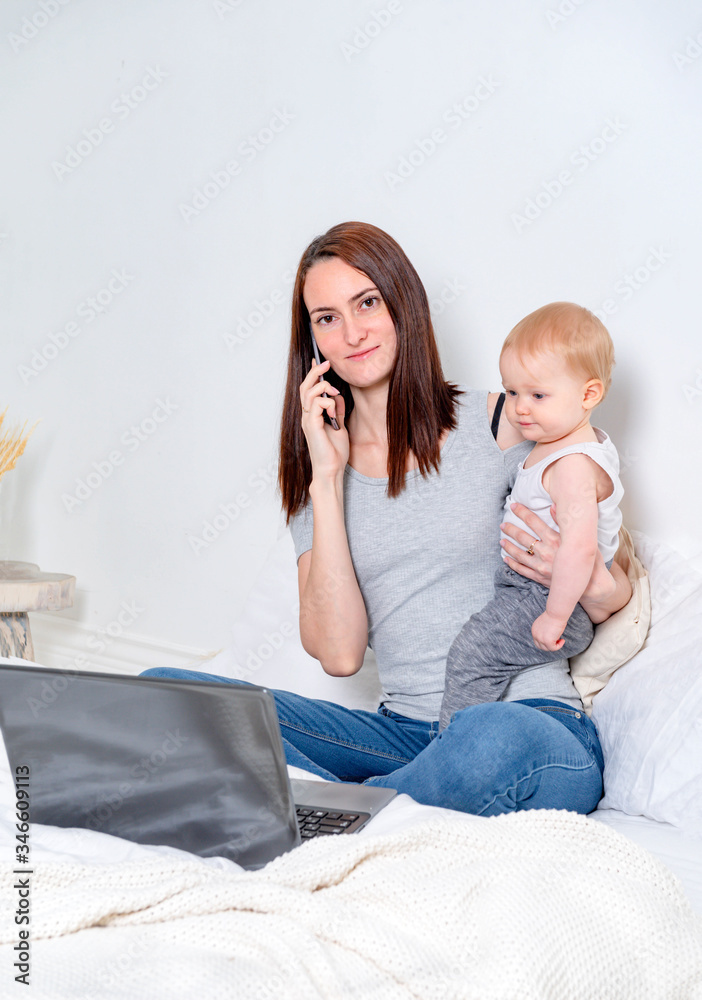 young mother sitting with a child on the bed and talking on the phone and working with a laptop, work at home,