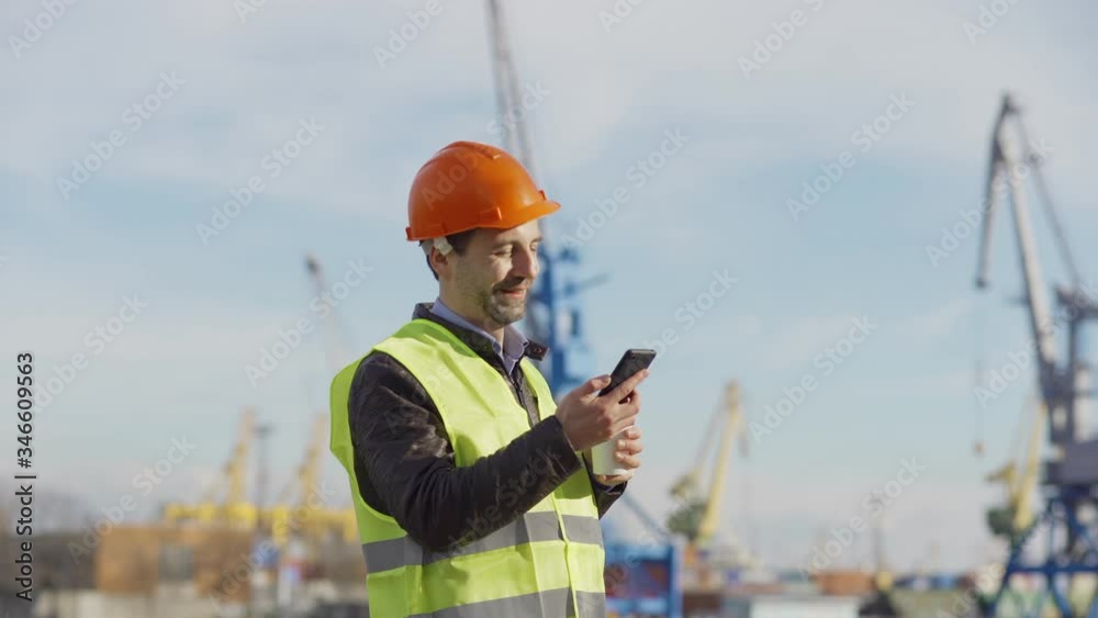 Side view of middle aged port engineer in helmet and safety vest standing at dockyard, drinking coffee and thinking. Man getting text message on cell phone, reading sms and smiling Side view of middle aged port engineer in helmet and safety vest standing at dockyard, drinking coffee and thinking. Man getting text message on cell phone, reading sms and smiling