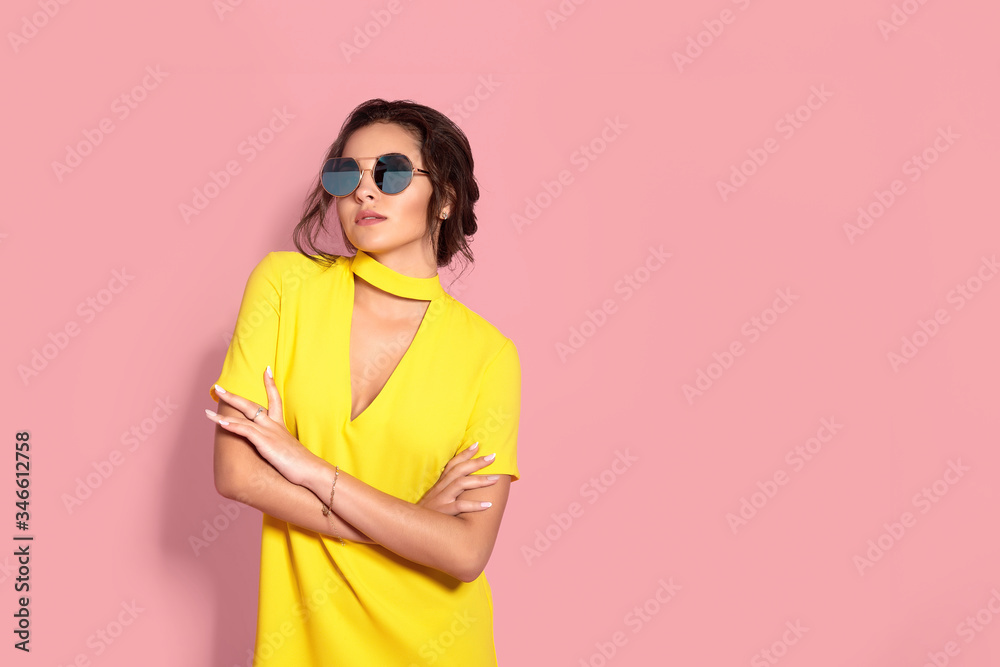 Beautiful girl wearing yellow dress and sunglasses posing on pink background in studio. Looking at camera.