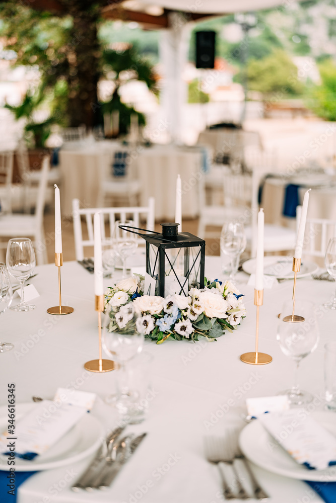 Wedding dinner table reception. A metal candlestick in the center of the table surrounded by floral composition, candles in gold candlesticks, white tables and plates with blue napkins