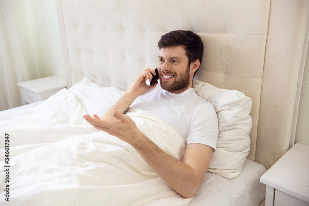 Man Talking on Phone in Bed. Morning at Home. Smilling Man Using Phone in Bed. Quarantine, Home Work, Medical Care. Man Talking on Phone in Bed. Morning at Home. Smilling Man Using Phone in Bed. Quarantine, Home Work, Medical Care.