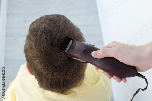 the hand holds an electric hair clipper and cuts the boy's long hair, barbershop at home, parent cuts hair while hairdressers are closed, stay home concept