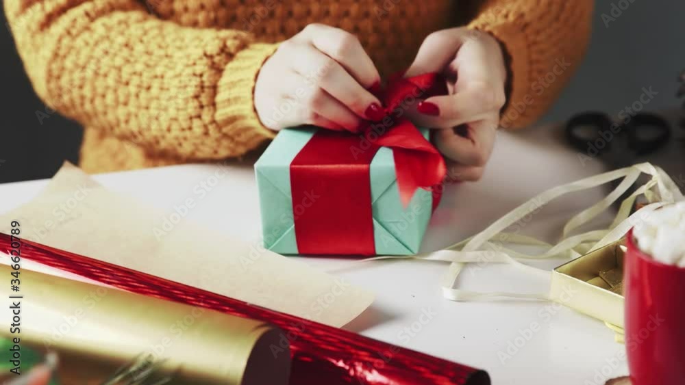 Female hands wrapping Christmas gift in blue paper and tying red ribbon
